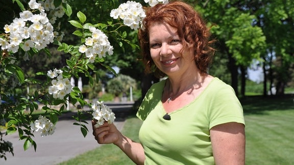 Eine Frau mit halblangen rot-braunen Haaren lächelt in die Kamera. Sie trägt ein hellgrünes T-Shirt und steht neben einer Pflanze mit weißen Blüten.