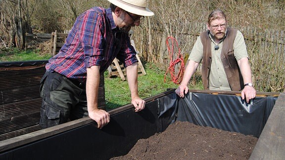 MDR Garten-Redakteur Jörg Heiß, ein Mann mit kariertem Hemd und geflochtenem Hut auf dem Kopf, und der Gemüseanbau-Experte von der Lehr- und Versuchsanstalt für Gartenbau in Erfurt, Martin Krumbein, stehen am Rand eines Hochbeetes mit Erde darin und stützen sich auf den hölzernen, mit schwarzer Folie ausgekleideten Rand auf.