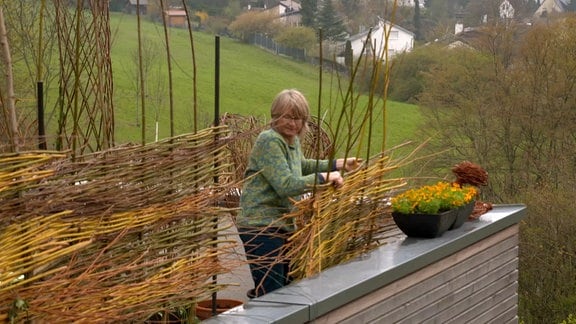 Eine Frau bringt einen Sichtschutz aus Weide an ihrem Balkon an. 