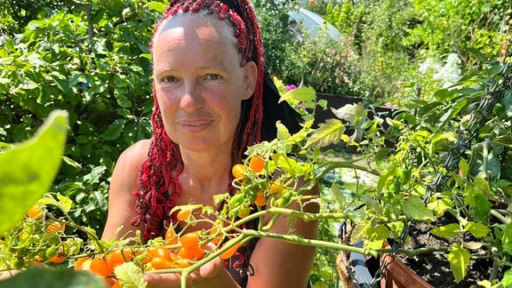 Eine Frau mit roten Rastalocken hält eine Tomatenpflanze voller gelber Tomaten in die Kamera.