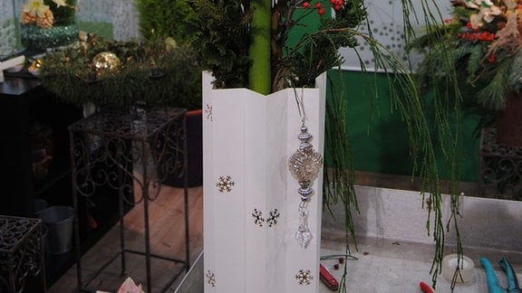 Ein Gesteck mit weiß blühendem Ritterstern, Früchten der Ebereschen an Ästen und Zweigen der Fadenzypresse in einer weißen Papiertüte.