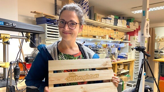 Reporterin Nadine Witt steht in einer Werkstatt und hält Teile einer Kiste mit eingebranntem Strebergärtner-Logo. 