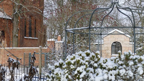 Garten mit einer Gartenlaube im Winter, im Hintergrund ein Ziegelgebäude
