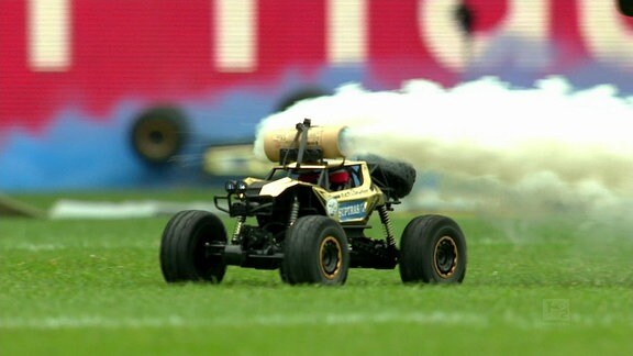 Ein elektrisches Spielzeugauto fährt über einen Fussballrasen und zieht eine Rauchwolke hinter sich her.