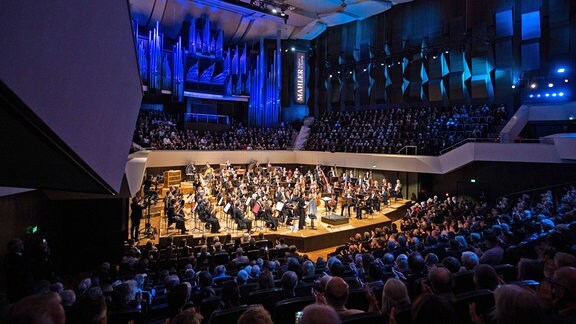 Blick ins Gewandhaus Leipzig: Der Saal ist voller Menschen, das Orchester steht im Zentrum der Bühne, die Wände dahinter sind blau angestrahlt.