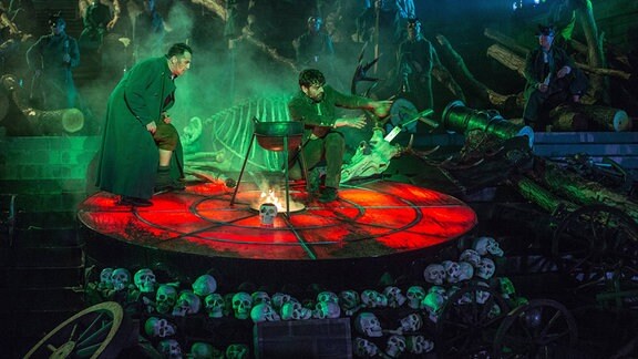 Zwei Personen mit langen Gewändern in einem grün-roten mystischen Bühnenbild, in der Mitte ein Dreibein-Kochtopf über Feuer, Totenköpfe und Skelette