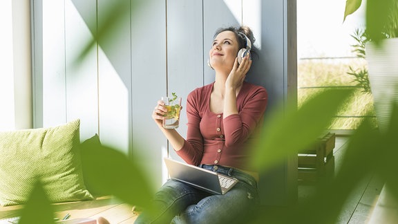 Eine weiße Frau hört mit Kopfhörern Musik, in der Hand ein gesundes Getränk, im Vordergrund unscharfe grüne Blätter
