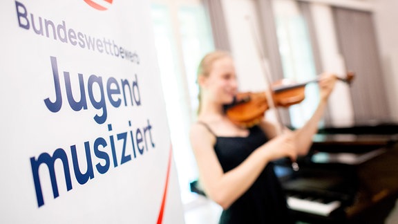 Die Aufschrift „Bundeswettbewerb Jugend musiziert" steht auf einem Plakat im Alten Landtag, während ein Mädchen ihre Geige spielt. Rund 2300 jugendliche Musikerinnen und Musiker nehmen an dem Bundeswettbewerb teil, der erstmals seit zwei Jahren wieder live und vor Publikum stattfindet.