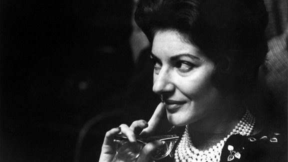 Schwarz-weiß-Porträt von Maria Callas, 1961: blick leicht zur rechten Seite, Hand zur Faus und Zeigefinger ausgestreckt auf Lippen, verschmitzter Blick, Augen-Make-Up, dunkle Haare, Kette mit vielen Perlenreihen