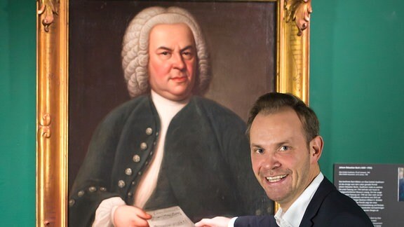 Mann im Anzug vor Gemälde von Johann Sebastian Bach. 