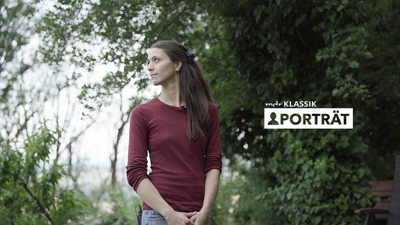 Junge Frau mit längeren, dunklen Haaren und rotem Oberteil und Jeans steht im Grünen, hält Hände zusammen, blickt zur Seite, im Hintergrund Bäume unscharf; Logo MDR KLASSIK-Porträt