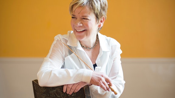 Porträt von Marin Alsop, seitlich auf einem Stuhl sitzend, zur Seite blickend mit freundlichem Lachen, vor gelbem Hintergrund.