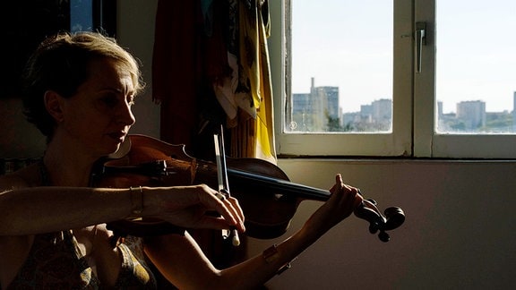 Die Bratschistin Maria Moscon aus Frankreichz sitzt daheim am Fenster und spielt ihr Instrument. Licht beleuchtet nur die Konturen sowie Licht im Fenster. 