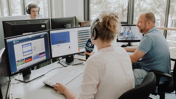 Ein Frau sitzt vor Computerbildschirmen und spricht in ein Mikrofon. Neben ihr sitzt ein Mann.