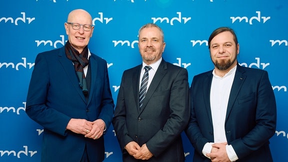 Drei Männer in Anzügen stehen vor einer blauen Wand mit dem Schriftzug MDR