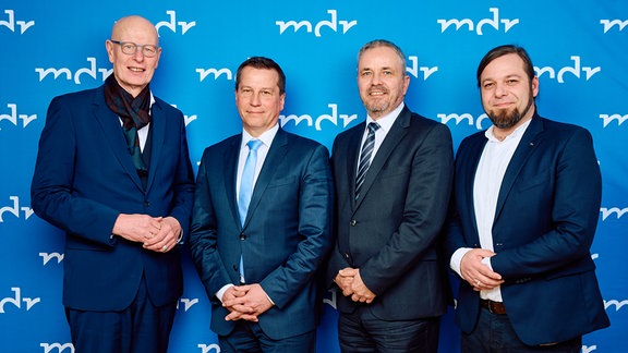 Vier Männer in Anzügen stehen vor einer blauen Wand mit dem Schriftzug MDR