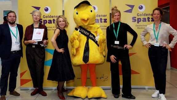 Vier Frauen und ein Mann stehen um das gelbe Plüsch-Maskottchen des Kinderfilmfestivals Golder Spatz.