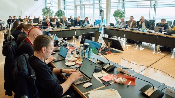 Mehrere Frauen und Männer sitzen an langen Konferenztischen. Vor ihnen stehen Laptops, Getränke und Gläser