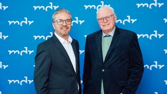 Zwei Männer in dunklen Anzügen und mit Brille stehen vor einer hellblauen Wand mit dem Logo mdr.