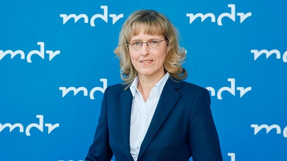 Eine Frau mit halblangen blonden Haaren und Brille mit dunkelblauem Blazer steht vor einer hellblauen Wand mit dem Logo mdr.