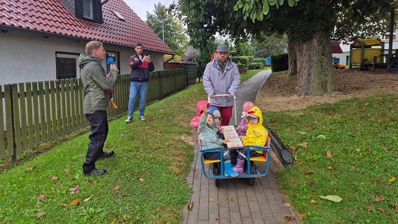 Ein Mann in grauer Regenjacke schiebt einen Wegen in dem mehrere Kinder sitzen