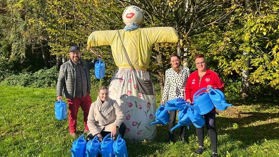 Vier Personen stehen neben einer Vogelscheuche mit blauen Kießkannen.