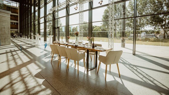 In einem sonnendurchfluteten Foyer steht eine minimalistisch gedeckte Tafel mit acht Stühlen, die lange Schatten wirft