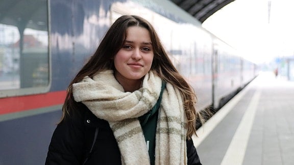 Ein junges Mädchen mit langen braunen Haaren steht auf einem Bahnsteig vor blauen Zugwaggons