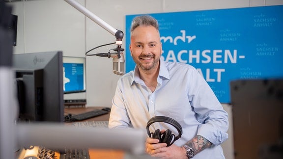 Junger Mann mit grauen Haare und hellblauem Hemd in einem Radiostudio mit Mikrofon