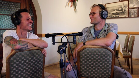 Zwei Männer mit Kopfhörern sitzen verkehrtherum auf Stühlen bei einem Interview