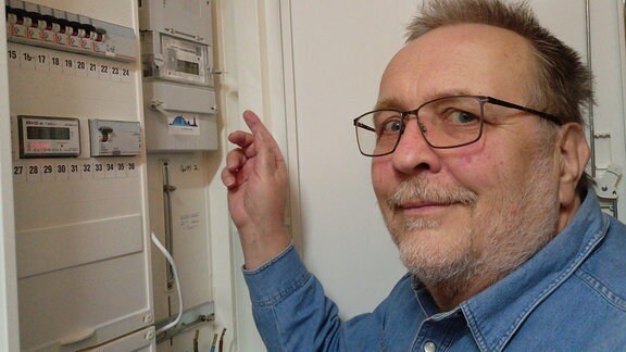 Mann in Jeanshemd, mit grauem Bart und Brille steht vor dem geöffneten Zähelrstand und zeigt auf den Smart Meter