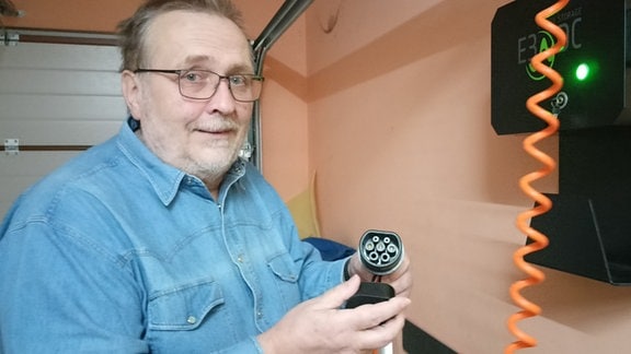 Mann in Jeanshemd, mit grauem Bart und Brille vor dem Stromanschluss seines Elektroautos in der Garage. In der Hand hält er einen großen Stecker
