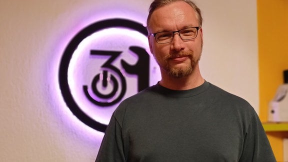 Stefan Haun von Netz39, ein junger Mann mit grauem T-Shirt und Brille vor einer Wand, im Hintergrund das Logo von Netz 39.