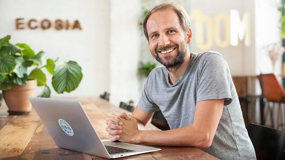 Junger Mann in grauem T-Shirt sitzt vor einem Laptop und lächelt in die Kamera.