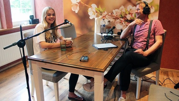 Blonde junge Frau und blonder Mann sitzen sich an einem Holztisch gegenüber mit Mikrofonen vor sich und einem Blütenbild an der Wand
