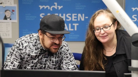 Zwei Personen stehen in einem Radiostudio – ein Mann mit schwarzer Kappe und eine Frau mit langen braunen Haaren