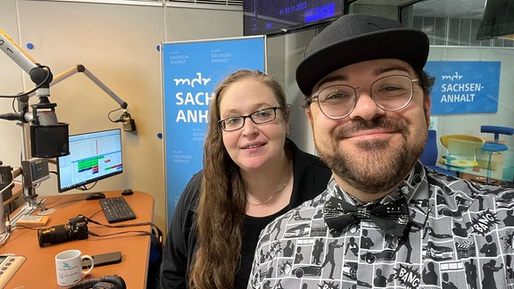 Zwei Personen stehen in einem Radiostudio – ein Mann mit schwarzer Kappe und eine Frau mit langen braunen Haaren