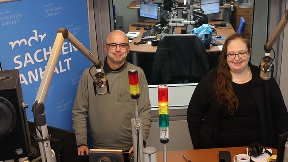 Zwei Personen stehen in einem Radiostudio – ein Mann mit schwarzer Kappe und eine Frau mit langen braunen Haaren