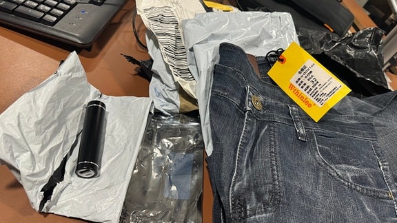 Eine Jeanshose mit dem Label "Wthinlee" und eine batteriebetriebene Powerbank liegen auf zerrissenen Versandpackungen.