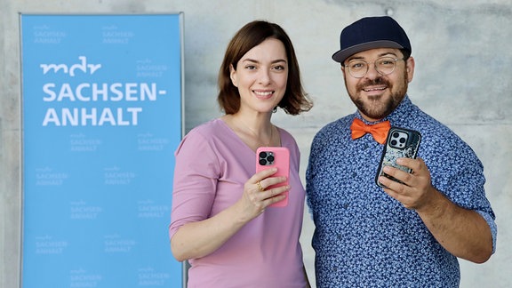 Eine Frau mit rosanem Oberteil und ein Mann mit blauem Hemd stehen vor einer Betonwand und lächeln