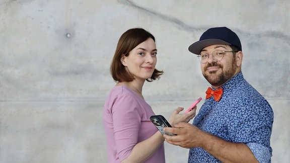 Eine Frau mit rosanem Oberteil und ein Mann mit blauem Hemd stehen vor einer Betonwand und lächeln