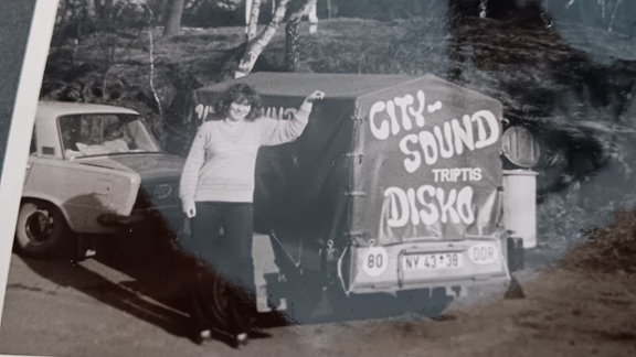 Auf einem historischen DDR-Foto in Schwarz-Weiß steht eine junge Frau an einen Auto-Anhänger gelehnt, auf dem für eine Disco geworben wird.