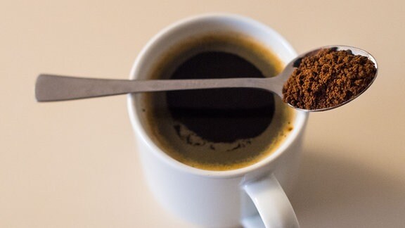 Ein Löffel mit Instant-Kaffeepulver liegt auf einer mit Instant-Kaffee gefüllten Kaffeetasse. 