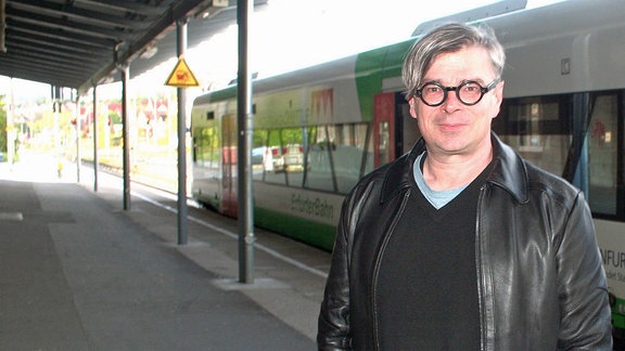 Ein Mann mit einer markanten schwarzen Brille steht vor einem Regionalzug am Bahnhof - es ist der tschechische SchriftstellerJaroslav Rudiš.
