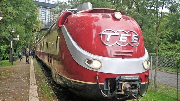 Ein Zug mit roter Lokomotive steht an einem Bahnsteig. Auf der Lokomotive steht in großen Buchstaben "TEE".