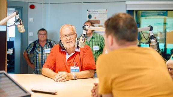 Zwei Personen sitzen sich an einem Tisch mit Radiotechnik gegenüber, im Hintergrund schauen weitere Menschen zu.