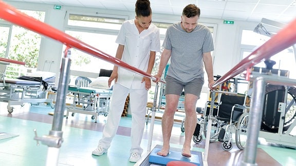 Ein Patient übt das Gehen unter Anleitung einer Therapeutin an einer Stange.