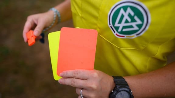 Eine Schiedsrichterin, steht mit roter und gelber Karte auf einem Rasen und hält in der anderen Hand eine Trillerpfeife. 