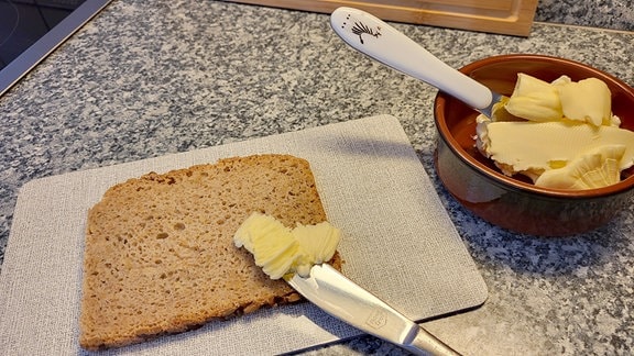 Neben einem braunen Keramikschälchen mit Butter liegt ein Brettchen mit einem Messer, das Butter auf ein Brot streicht.