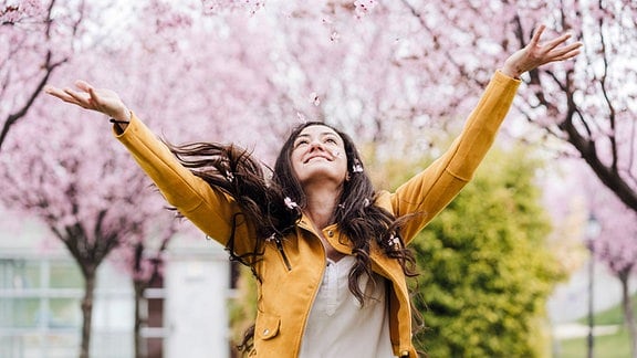 Junge Frau freut sich, mit einer leichten Übergangsjacke bekleidet, über die Kirschblüte im Frühling.